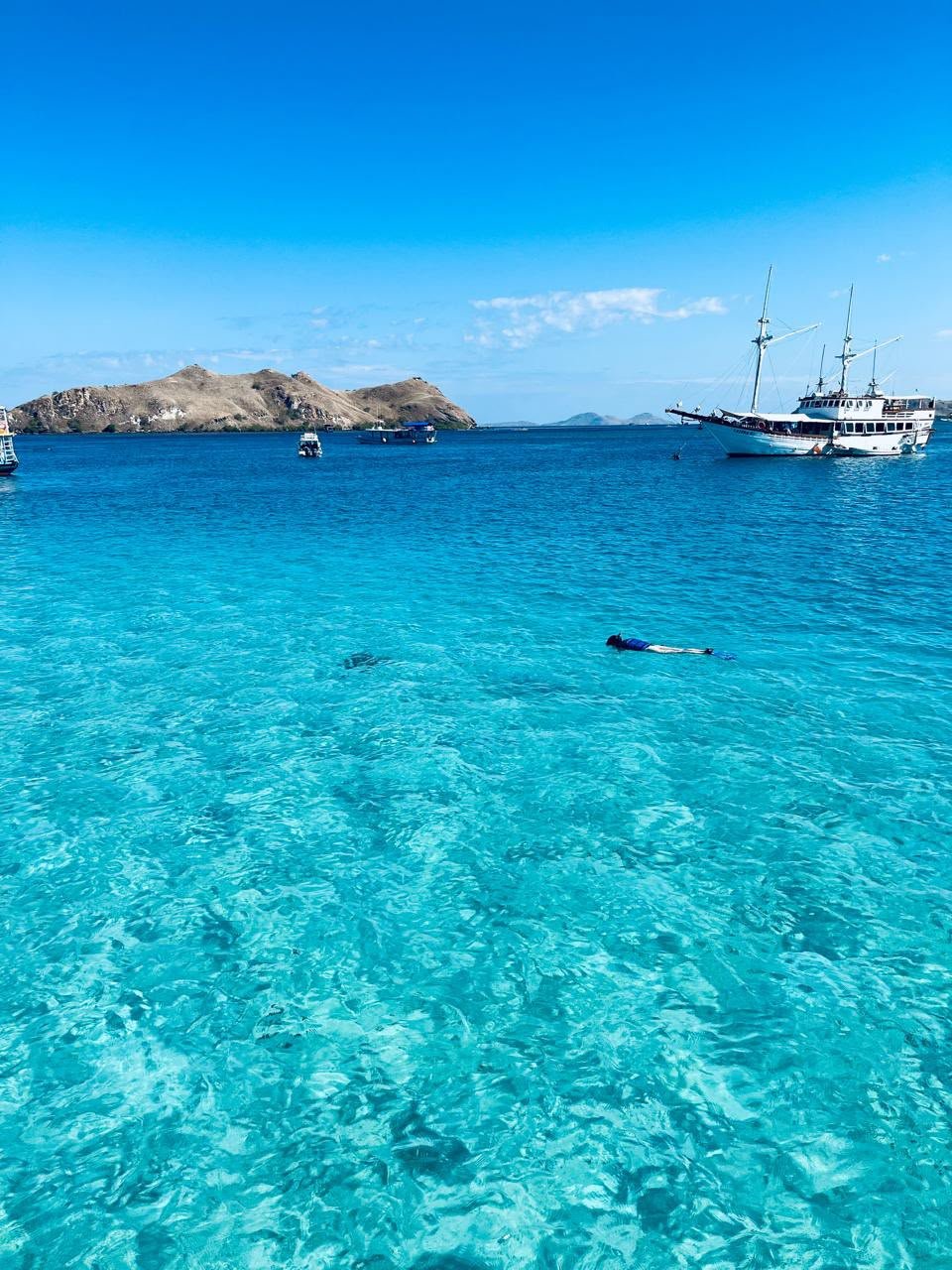 Snorkeller floating above clear turquoise shallows with a phinisi boat anchored offshore in Komodo National Park