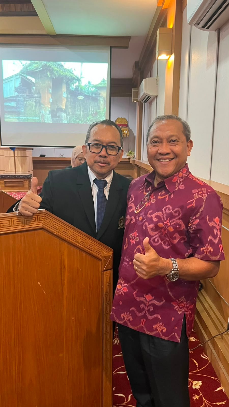 Dr. Ketut Jaman and Dr. Trisno Nugroho beside the lectern, with a photograph of Penglipuran Tourism Village on the presentation screen behind them